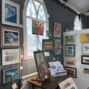 Colourful paintings and original artworks displayed beside the arched windows of the Tin Tabernacle at hART 2025.