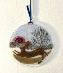 Circular fused glass artwork by Caroline Raffan depicting a running hare beneath a stylised tree and colourful sky.