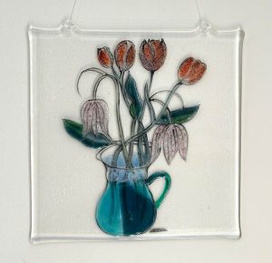 Fused glass artwork by Caroline Raffan featuring tulips and delicate flowers arranged in a turquoise-blue jug.