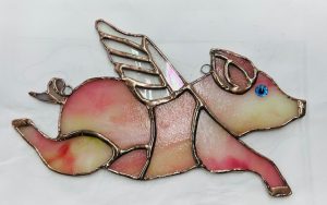 Stained glass flying pig by Yolanda Houston in soft pink and cream glass with copper-toned outlines and a bright blue eye.